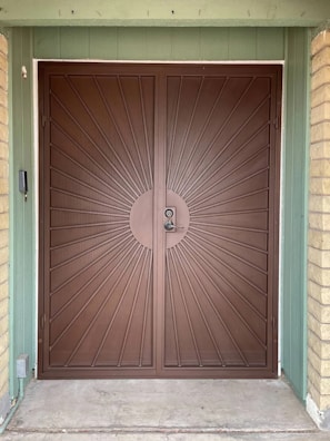 A large, double brown metal door with a sunburst design pattern originating from a central circular point where the door handles are located. The door is framed by a green wooden structure and two beige brick columns on each side.