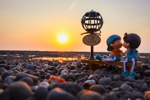 A pair of figurines representing a couple are sitting on a small bench on a pebbly beach, with a sunset in the background. The figures are kissing, and in the foreground is a decorative lamp with a sign that reads 'Love'. The warm glow of the setting sun creates a romantic and serene atmosphere.
