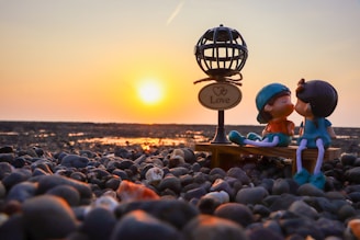 A pair of figurines representing a couple are sitting on a small bench on a pebbly beach, with a sunset in the background. The figures are kissing, and in the foreground is a decorative lamp with a sign that reads 'Love'. The warm glow of the setting sun creates a romantic and serene atmosphere.