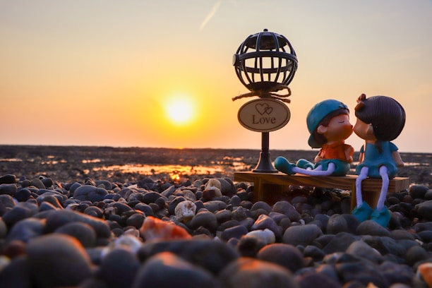 A pair of figurines representing a couple are sitting on a small bench on a pebbly beach, with a sunset in the background. The figures are kissing, and in the foreground is a decorative lamp with a sign that reads 'Love'. The warm glow of the setting sun creates a romantic and serene atmosphere.