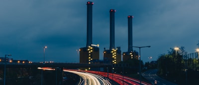 Three tall chimneys are part of an industrial power plant, surrounded by evening twilight. Bright lights illuminate the structures, contrasting with the darkening sky. In the foreground, light trails from vehicles create dynamic lines, indicating movement on the highway nearby.