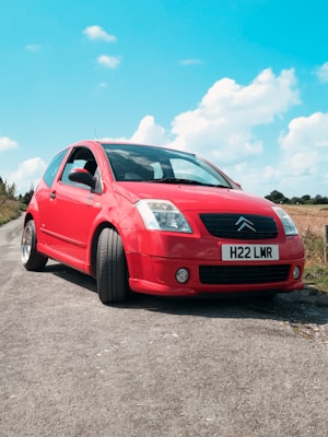 A bright red car is parked on a rural road under a blue sky with scattered clouds. The car is a Citroën with a license plate that reads H22 LMR. The road is bordered by grass and some bushes.