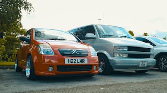 Two cars are parked in an open parking lot. The car on the left is a bright orange Citroën, while the car on the right is a silver van. The cars are parked next to each other on a paved surface with some greenery visible in the background.