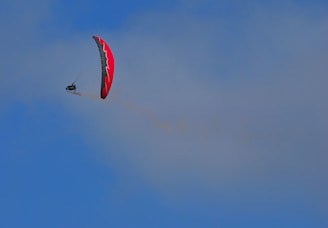 A paraglider with a red and black canopy is soaring high against a clear blue sky. The paraglider is slightly tilting, creating a dynamic sense of motion and freedom in the air.