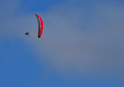A paraglider with a red and black canopy is soaring high against a clear blue sky. The paraglider is slightly tilting, creating a dynamic sense of motion and freedom in the air.