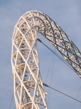 A close-up view of a large architectural structure featuring a complex, white lattice framework made of intersecting beams. The structure is set against a clear blue sky, with cables providing additional support. The design is both intricate and minimal, showcasing engineering precision.