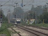 A train is approaching, traveling down the railway tracks under a network of overhead electric wires. The tracks are flanked by greenery, with visible trees and foliage. There is a person standing near the tracks, possibly railway staff. Signal lights and various signs are positioned along the tracks, and in the background, a small structure is visible, possibly part of a railway station.