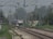 A train is approaching, traveling down the railway tracks under a network of overhead electric wires. The tracks are flanked by greenery, with visible trees and foliage. There is a person standing near the tracks, possibly railway staff. Signal lights and various signs are positioned along the tracks, and in the background, a small structure is visible, possibly part of a railway station.