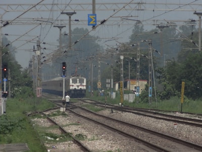 A train is approaching, traveling down the railway tracks under a network of overhead electric wires. The tracks are flanked by greenery, with visible trees and foliage. There is a person standing near the tracks, possibly railway staff. Signal lights and various signs are positioned along the tracks, and in the background, a small structure is visible, possibly part of a railway station.