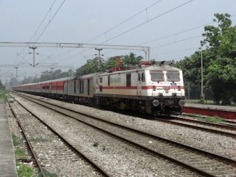 A passenger train travels along railway tracks, featuring a white and red locomotive with the text 'Indian Railways' on its side. The train is moving away from a station platform lined with overhead electrical wires and surrounded by green trees. The sky is overcast, adding a calm ambiance to the setting.