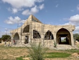 A large building under construction with a distinct arched design. The structure is surrounded by scaffolding, indicating ongoing work. The building is set against a backdrop of a partly cloudy sky, and the ground is predominantly grassy with some patches of dirt.