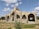 A large building under construction with a distinct arched design. The structure is surrounded by scaffolding, indicating ongoing work. The building is set against a backdrop of a partly cloudy sky, and the ground is predominantly grassy with some patches of dirt.