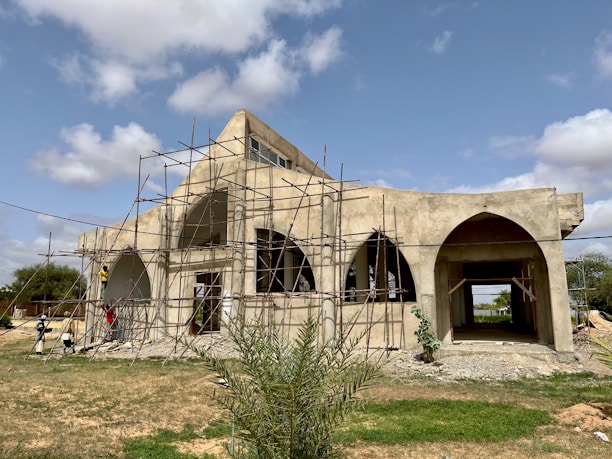 A large building under construction with a distinct arched design. The structure is surrounded by scaffolding, indicating ongoing work. The building is set against a backdrop of a partly cloudy sky, and the ground is predominantly grassy with some patches of dirt.