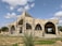 A large building under construction with a distinct arched design. The structure is surrounded by scaffolding, indicating ongoing work. The building is set against a backdrop of a partly cloudy sky, and the ground is predominantly grassy with some patches of dirt.