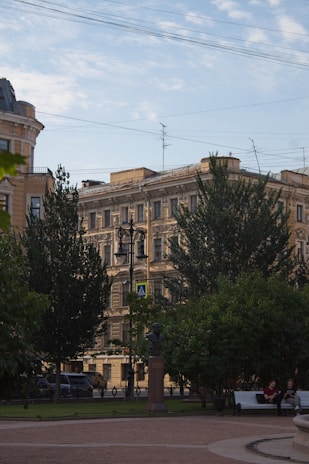 A city scene featuring a historical building with detailed architectural features surrounded by trees. In front of the building is a bronze bust on a pedestal situated in a small park area. Two people are seated on a bench enjoying the scenery, and a classic streetlamp is prominently displayed.