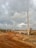 A dirt road winds through a rugged landscape under a cloudy sky. There are several electricity poles alongside the road. A few people can be seen in the distance, standing near some construction materials. In the background, there are piles of earth and sparse greenery, indicating a possible construction site.