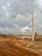 A dirt road winds through a rugged landscape under a cloudy sky. There are several electricity poles alongside the road. A few people can be seen in the distance, standing near some construction materials. In the background, there are piles of earth and sparse greenery, indicating a possible construction site.