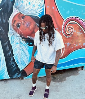 A person with dreadlocks, wearing a white shirt, dark shorts, and sunglasses stands in front of a vibrant mural. The mural depicts a stylized female figure with intricate details and a rich color palette, including elements of an octopus with swirling tentacles.