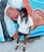 A person with dreadlocks, wearing a white shirt, dark shorts, and sunglasses stands in front of a vibrant mural. The mural depicts a stylized female figure with intricate details and a rich color palette, including elements of an octopus with swirling tentacles.