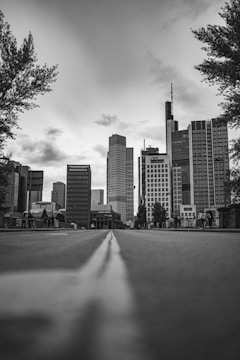 A grayscale photograph of a cityscape featuring modern skyscrapers lining a broad, deserted avenue. Trees frame the image on either side, and the perspective is from a low angle close to the ground, with the street's center line leading towards the tall buildings. A few people are visible in the distance, adding a sense of scale.