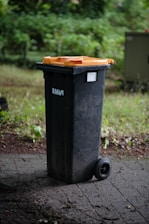 A black wheeled trash bin with an orange lid stands on a paved surface surrounded by greenery. The surroundings are slightly blurred, indicating a shallow depth of field.