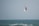 A person is engaged in kitesurfing, skillfully maneuvering high above the ocean waves. They are holding onto a kite that provides lift and control as they soar through the air. The ocean beneath them appears rough with small waves, and the sky is a plain light blue color.