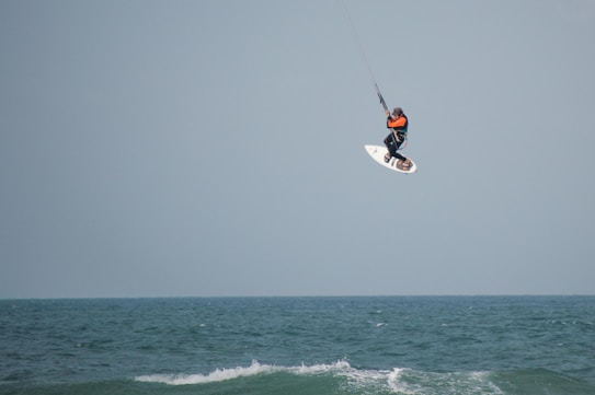A person is engaged in kitesurfing, skillfully maneuvering high above the ocean waves. They are holding onto a kite that provides lift and control as they soar through the air. The ocean beneath them appears rough with small waves, and the sky is a plain light blue color.