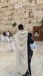 A man in traditional Jewish attire stands in front of a large stone wall, which is the Western Wall. He is wearing a kippah and a tallit. Several white plastic chairs are scattered around the area, and other people are visible near the wall, some engaged in prayer. A wooden cabinet-like structure is positioned against the wall to the right.