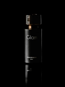 A sleek, dark bottle labeled 'Glam' with gold accents is centered against a black background. The reflective surface beneath adds a sense of elegance and luxury to the presentation.