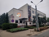 A large, rectangular building with a modern design featuring clean lines and a smooth facade. The building is adorned with a logo reading 'radio LODZ' on its side. There is a row of windows along the upper floors, and the entrance is marked by a small awning. Signs indicating no stopping and no entry for certain vehicles are visible nearby. Surrounding the building are green shrubs and trees lining the sidewalk.