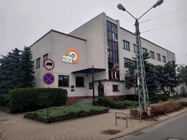 A large, rectangular building with a modern design featuring clean lines and a smooth facade. The building is adorned with a logo reading 'radio LODZ' on its side. There is a row of windows along the upper floors, and the entrance is marked by a small awning. Signs indicating no stopping and no entry for certain vehicles are visible nearby. Surrounding the building are green shrubs and trees lining the sidewalk.