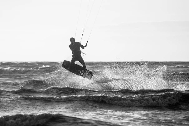 A person is engaged in kitesurfing, performing a dynamic maneuver above the waves with spray flying through the air. The silhouette is captured against the backdrop of the ocean, highlighting the movement and energy of the sport.