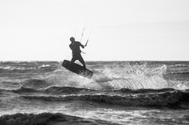 A person is engaged in kitesurfing, performing a dynamic maneuver above the waves with spray flying through the air. The silhouette is captured against the backdrop of the ocean, highlighting the movement and energy of the sport.