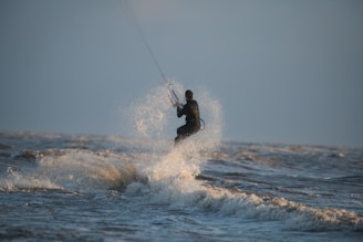 A person is engaging in kite surfing, skillfully maneuvering over the ocean waves with a kite in hand. The water sprays around them, creating an exhilarating sense of motion and adventure against the backdrop of a clear sky.