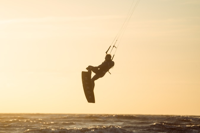A silhouetted kite surfer is captured mid-air against a backdrop of a sunset-lit sky and ocean, creating a dramatic and dynamic scene. The surfer is elevated on the left side of the image with the kite strings visible, and the water appears slightly turbulent.