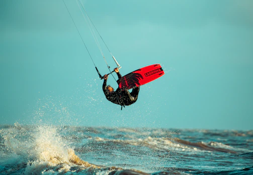 A person is performing a kite surfing maneuver above the ocean, holding onto a kite bar. The kiteboard is red and visible against the backdrop of the sky and ocean. Waves are crashing below, and the lighting suggests a sunny day.