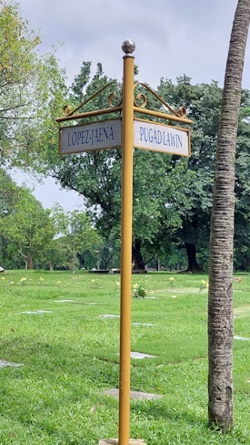 A street sign post stands prominently in a lush green park, displaying two street names: Lopez-Jaena and Pugad Lawin. The surrounding area is filled with verdant grass, scattered trees, and a few stone slabs on the ground. The sky appears overcast with light cloud cover, and there is a tall palm tree in the background.