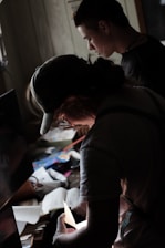 Two individuals are focused on examining various papers and items on a cluttered desk. The lighting is dim, casting shadows across the scene while illuminating the papers being handled. The atmosphere suggests a sense of concentration and attention to detail.