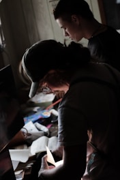 Two individuals are focused on examining various papers and items on a cluttered desk. The lighting is dim, casting shadows across the scene while illuminating the papers being handled. The atmosphere suggests a sense of concentration and attention to detail.