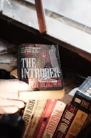 A gloved hand holds a book titled 'The Intruder' near a dusty, cracked window. Several other books are visible on the wooden shelf below, including titles by Arthur Hailey and John le Carré. The scene suggests an old or abandoned setting.