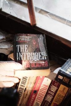 A gloved hand holds a book titled 'The Intruder' near a dusty, cracked window. Several other books are visible on the wooden shelf below, including titles by Arthur Hailey and John le Carré. The scene suggests an old or abandoned setting.