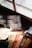 A gloved hand holds a book titled 'The Intruder' near a dusty, cracked window. Several other books are visible on the wooden shelf below, including titles by Arthur Hailey and John le Carré. The scene suggests an old or abandoned setting.