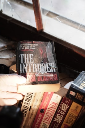 A gloved hand holds a book titled 'The Intruder' near a dusty, cracked window. Several other books are visible on the wooden shelf below, including titles by Arthur Hailey and John le Carré. The scene suggests an old or abandoned setting.