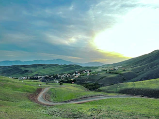 Lush green hills in Albania with a winding road leading toward a sunlit village nestled in the valley.