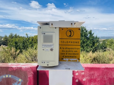 A public emergency phone is mounted on a red and white painted platform outdoors, with a backdrop of greenery and a cloudy blue sky. The phone unit has a speaker and a button, with a yellow sign next to it displaying instructions in French for use in case of an anomaly.