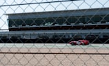 A red race car is speeding on a race track in front of a modern building with large windows. The image is taken through a chain-link fence, adding a textured overlay to the scene. There is a gravel path in the foreground, and the sky is clear.