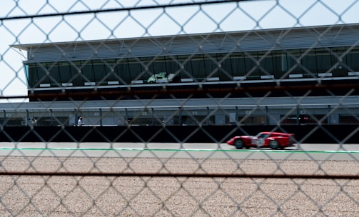 A red race car is speeding on a race track in front of a modern building with large windows. The image is taken through a chain-link fence, adding a textured overlay to the scene. There is a gravel path in the foreground, and the sky is clear.