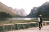 A scenic view of a tranquil lake surrounded by tall mountains and a dense forest. A person with a backpack is walking on a path beside the lake, holding a yellow umbrella. The path is bordered by a wooden railing, and a lush grassy area with wildflowers is visible in the foreground.