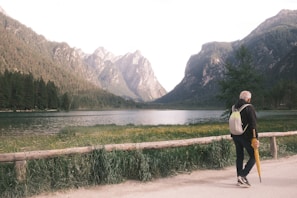 A scenic view of a tranquil lake surrounded by tall mountains and a dense forest. A person with a backpack is walking on a path beside the lake, holding a yellow umbrella. The path is bordered by a wooden railing, and a lush grassy area with wildflowers is visible in the foreground.