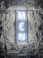 A neon sign glowing brightly against a textured stone wall, displaying the word 'MEN' in vertical alignment with white light. The illumination casts a soft glow around the sign, highlighting the rough surface of the wall.